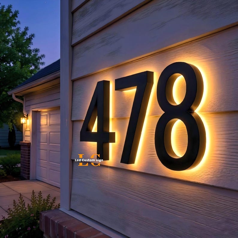 May include: Black illuminated house numbers 478 mounted on a light-colored wooden wall. The numbers are backlit with a warm, golden glow. The text "Led Custom sign" is visible in the lower left corner. The scene is set at dusk.