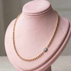 Può includere: Una collana dorata con perline con un piccolo pendente ovale con un gruppo di piccole pietre trasparenti. La collana è esposta su un busto di gioielli rosa.