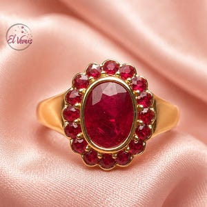 May include: A gold ring featuring a large oval-cut red gemstone at the centre, surrounded by a halo of smaller round-cut red gemstones. The metal band is smooth and polished. The text El Venus is visible in the upper left corner of the image.