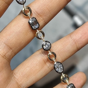 Puede incluir: Una pulsera con eslabones alternos de anillos de oro pulido y engastes de metal oscuro. Los engastes oscuros sostienen piedras claras facetadas en formas ovaladas y rectangulares. Pequeñas piedras brillantes están incrustadas en los conectores de oro.