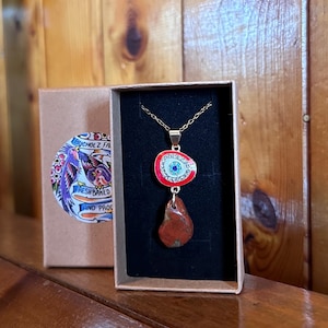 May include: A gold-colored chain necklace with a pendant featuring a red and white circular design with a blue center, and a teardrop-shaped, reddish-brown stone. The necklace is displayed in a black box, with a brown box and a sticker with text visible in the background.
