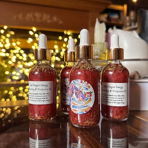 May include: Several small glass bottles with gold-colored tops and white droppers. The bottles are filled with a red liquid and herbs. The labels read "Red Dragon Energy Protecting & Protection Oil" and "Fresh Baked Goods".