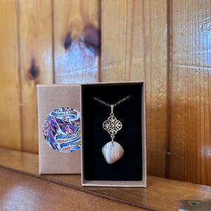 May include: A gold-colored pendant necklace with a patterned design and a stone pendant. The necklace is displayed in a black box. A cardboard box with a colorful sticker is to the left of the necklace.