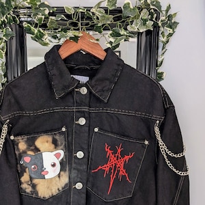 Op de afbeelding: Zwart denim jack met een cartoon hond patch op de ene zak en een rood geborduurd symbool op de andere. Het jack heeft zilveren kettingen aan de mouwen en het woord "NEXGEN" geborduurd op de ene mouw.