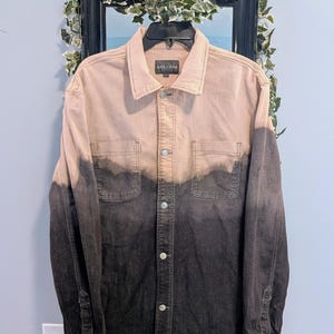 May include: A two-tone denim jacket with a light tan upper half and a dark gray lower half. The jacket has a button-down front, two chest pockets, and buttoned cuffs. The brand name "SLATE & STONE" is visible on the label.