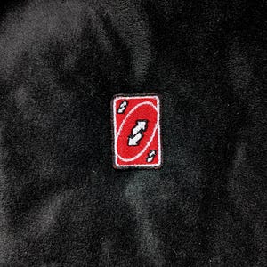 May include: A rectangular patch featuring a red background with a white arrow symbol and white accents. The patch is a reverse card from the popular card game, Uno. The patch is set against a dark, textured background.