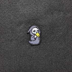 May include: Embroidered patch depicting a cartoon Grim Reaper in a grey robe, holding a yellow object. The patch has a white skull face and is set against a dark grey background. Suitable for jackets, bags, or other fabric items.
