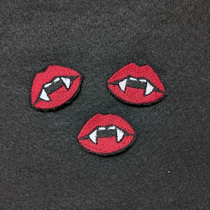 May include: Three embroidered patches of red lips with white fangs and black outlines. The patches are arranged in a triangular pattern against a dark gray fabric background. The patches are likely for clothing or accessories.