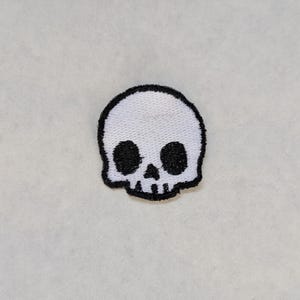 Könnte beinhalten: Ein kleiner, weißer Totenkopf-Aufnäher mit schwarzen Augenhöhlen und schwarzem Umriss. Der gestickte Aufnäher hat ein minimalistisches Design. Der Totenkopf ist ein beliebtes Symbol in der Gothic- und Alternativmode.