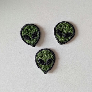 Puede incluir: Tres pequeños parches bordados con forma de cabeza de alienígena. Los parches presentan una base de hilo verde vibrante con detalles en hilo negro para los grandes ojos almendrados y un borde negro grueso que perfila la forma de la cabeza.