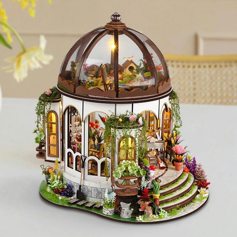 May include: A miniature greenhouse model with a domed roof and intricate details. The structure features a white exterior with brown accents, arched windows, and a variety of colorful flowers and greenery. The interior is illuminated, showcasing a detailed garden scene.