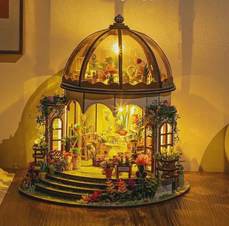 May include: A miniature greenhouse model with a domed glass roof and multiple levels. The interior is illuminated, showcasing tiny plants, furniture, and decorative elements. The exterior features flower pots and greenery, creating a charming, detailed scene.