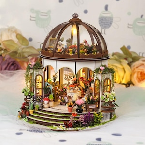 May include: A miniature wooden greenhouse with a glass dome, filled with tiny plants and flowers. The structure has a white base, brown trim, and is illuminated with warm lighting. The scene includes miniature furniture and decorative elements.