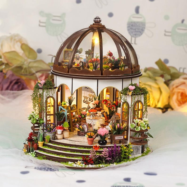 May include: A miniature wooden greenhouse with a glass dome, filled with tiny plants and flowers. The structure has a white base, brown trim, and is illuminated with warm lighting. The scene includes miniature furniture and decorative elements.