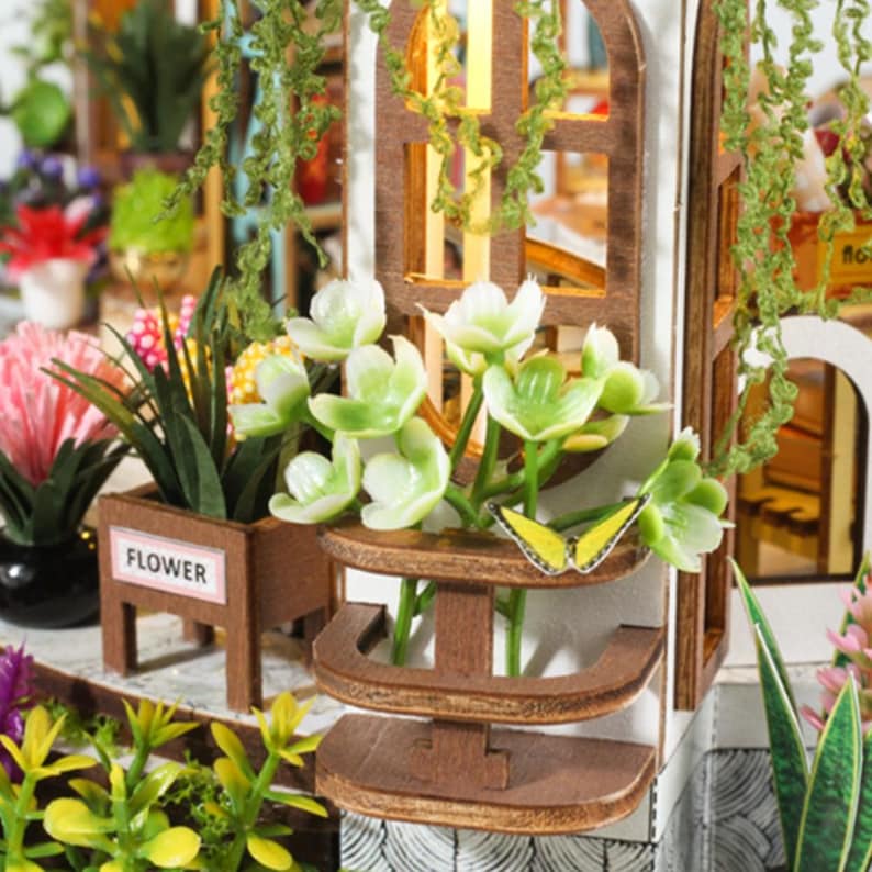 May include: A miniature wooden dollhouse with a flower shop theme. The facade features a brown window frame, white walls, and a small wooden shelf with green and white flowers. A yellow butterfly decorates the shelf.