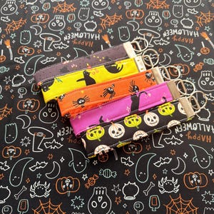 May include: Four fabric wristlet keychains with silver-tone metal hardware. The wristlets feature Halloween-themed patterns including black cats on dark gray, black cats on yellow, spiders on orange, and black cats on purple, plus a pumpkin print.