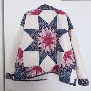 May include: A quilted jacket featuring a star pattern in shades of deep blue, magenta, and white. The garment displays a floral print on the blue and magenta sections, contrasted with solid white panels, long sleeves, and a collared neckline.