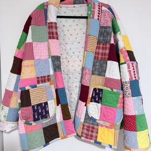 May include: A patchwork jacket with a variety of colorful fabric squares. The jacket has two front pockets and a floral patterned lining. The jacket is hanging on a black hanger against a white background.