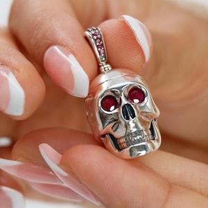May include: A silver skull pendant with red gemstone eyes and a gemstone-set bale. The skull has detailed features. The pendant is held by a hand with manicured nails.