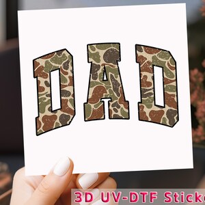 May include: A white square sticker with the word "DAD" in large, block letters. The letters are filled with a camouflage pattern in shades of brown, green, and tan, outlined in black. The text "3D UV-DTF Sticker" is at the bottom.