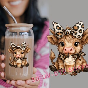 May include: A clear glass tumbler with a bamboo lid and a stainless steel straw. The tumbler features a cartoon cow with a leopard print bow and scarf. The cow is drinking from a coffee drink. The tumbler is filled with a brown liquid.