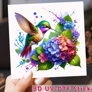May include: A colorful 3D UV-DTF sticker featuring a hummingbird in flight with a vibrant green, blue, and red color scheme. The hummingbird is surrounded by a cluster of pink, blue, and purple hydrangea flowers with green leaves and colorful paint splatters.