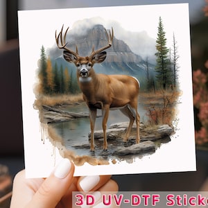May include: A 3D UV-DTF sticker featuring a watercolor illustration of a white-tailed deer standing in a forest stream. The deer has large antlers and is surrounded by trees and mountains.