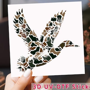 May include: A 3D UV-DTF sticker featuring a camouflage duck in flight. The duck is outlined in black and has a brown and green camouflage pattern.