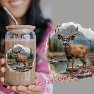 May include: A clear glass tumbler with a brown liquid inside. The tumbler has a straw sticking out of it. The tumbler has a white deer with antlers in a forest scene printed on it. The scene includes a mountain, trees, and a river.
