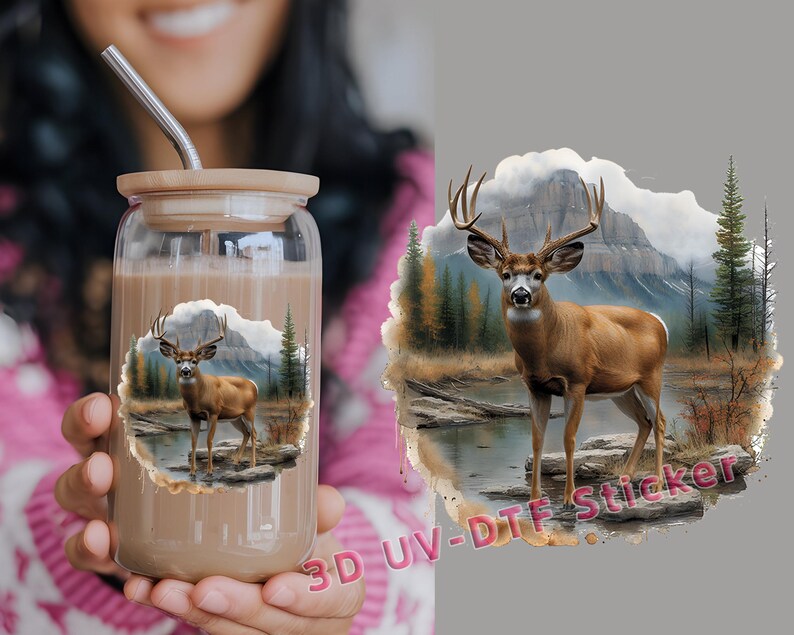 May include: A clear glass tumbler with a brown liquid inside. The tumbler has a straw sticking out of it. The tumbler has a white deer with antlers in a forest scene printed on it. The scene includes a mountain, trees, and a river.
