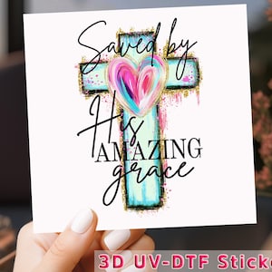 May include: A white square sticker featuring a cross with a heart in the center. The cross is light blue with gold glitter accents. The heart is a mix of pink, blue, and purple. The text reads "Saved by His Amazing Grace."