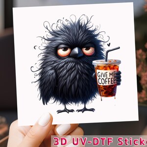 May include: A whimsical illustration of a grumpy, black, fluffy bird holding an iced coffee. The coffee cup reads "GIVE ME COFFEE". The bird has large, expressive eyes and a slightly annoyed expression. A black straw is in the coffee.