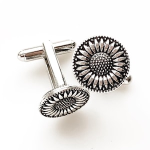 May include: A pair of silver cufflinks with a sunflower design. The cufflinks are round and have a detailed sunflower design with a textured center.