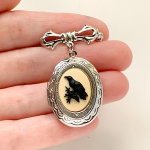 May include: A silver-toned oval locket brooch featuring a bow-shaped pin at the top. The locket has an ornate scalloped border and a central cream-colored inlay depicting a black raven perched on a branch. The metal has a polished finish.