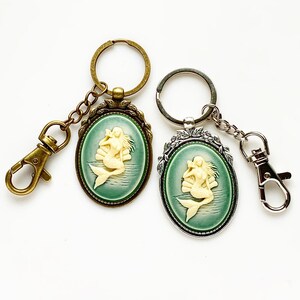 May include: Two keychains with oval-shaped charms featuring a white cameo of a mermaid with a green background. One keychain is bronze and the other is silver.