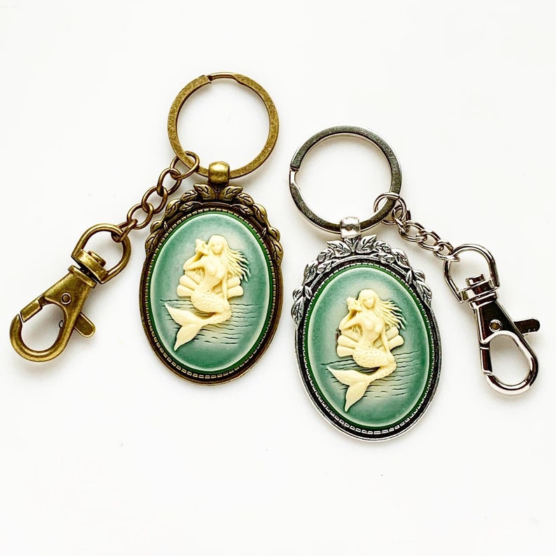 May include: Two keychains with oval-shaped charms featuring a white cameo of a mermaid with a green background. One keychain is bronze and the other is silver.