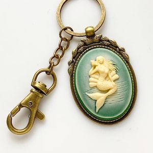 May include: A bronze key chain with a green cameo featuring a white mermaid figurine. The mermaid is sitting on a seashell and holding a seashell to her ear.
