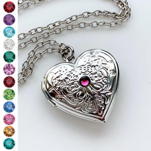 May include: A silver heart-shaped locket necklace with a pink rhinestone in the center. The locket has an ornate design and is on a silver chain.