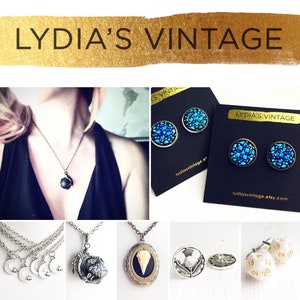 May include: A collection of jewelry featuring a variety of styles, including a silver necklace with a black stone pendant, a pair of blue crystal earrings, a silver necklace with a moon charm, a silver necklace with a dice charm, a silver necklace with a locket charm, and a silver pin with a thistle design. The jewelry is displayed on a white background with the text "LYDIA'S VINTAGE" on the top.
