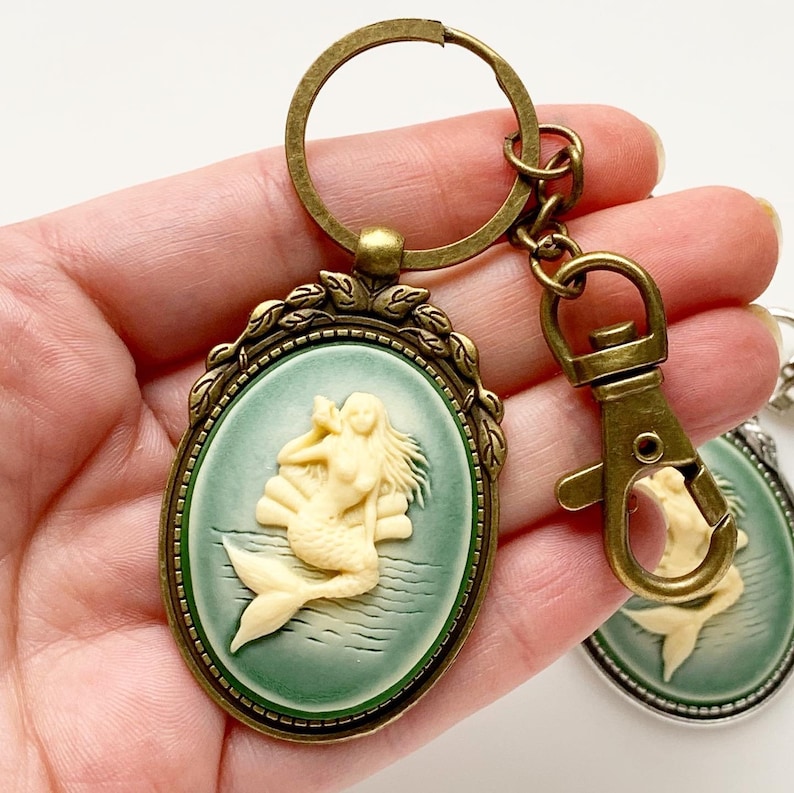 May include: A bronze key chain with a green cameo featuring a white mermaid figurine. The cameo is surrounded by a decorative bronze frame with leaf accents.