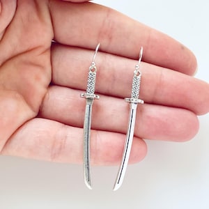 May include: A pair of silver earrings shaped like swords. The swords have a detailed handle and a long, thin blade.