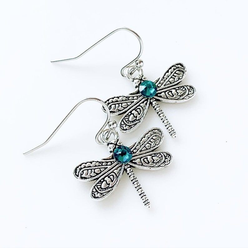 May include: Silver dragonfly earrings with teal blue crystal accents. The earrings have a detailed design and dangle from silver hooks.