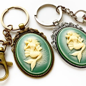 May include: Two keychains with a cameo of a mermaid in a green and white oval setting. One keychain is silver and the other is bronze.