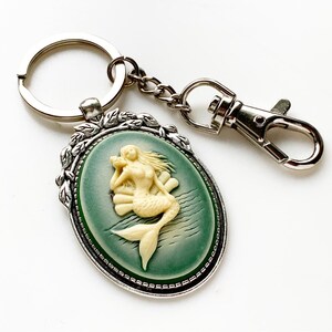 May include: A silver key chain with a green cameo featuring a white mermaid figurine. The cameo is surrounded by a silver frame with leaf accents.