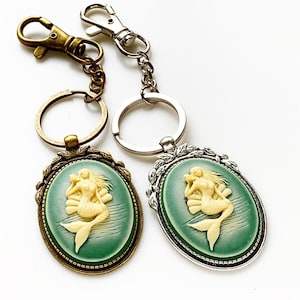 May include: Two keychains with a green and white cameo of a mermaid. One keychain is silver and the other is bronze.