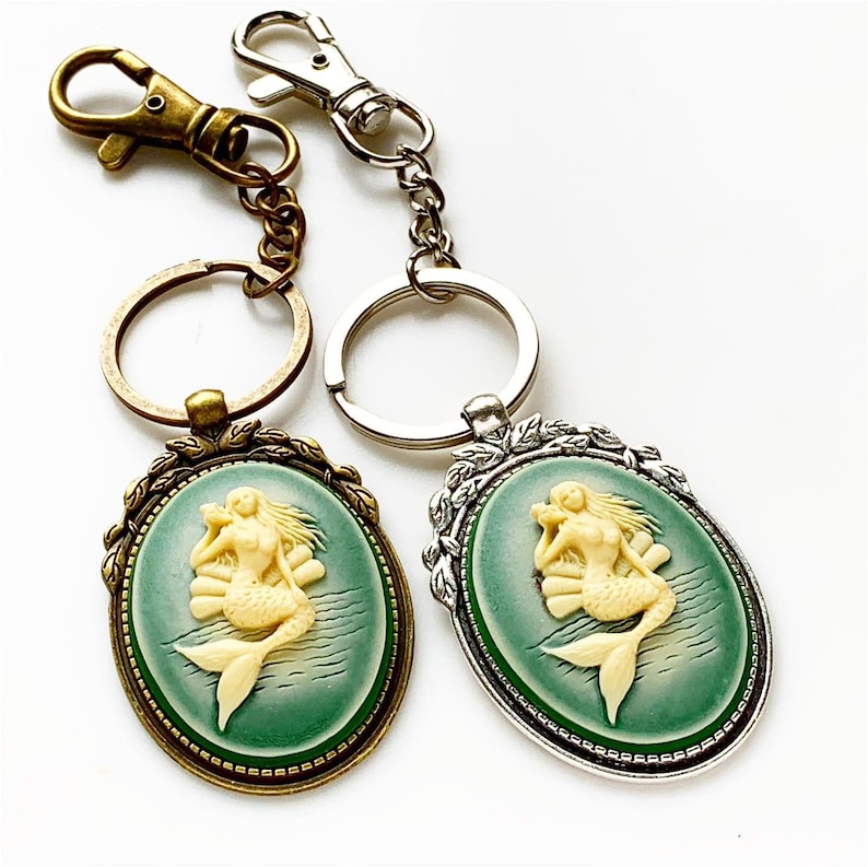 May include: Two keychains with a green and white cameo of a mermaid. One keychain is silver and the other is bronze.