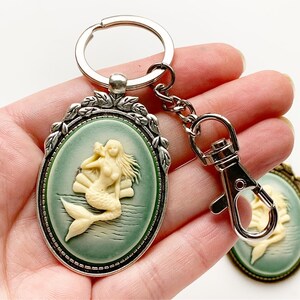 May include: A silver key chain with a green oval cameo featuring a white mermaid figurine. The cameo is surrounded by a silver frame with leaf accents.