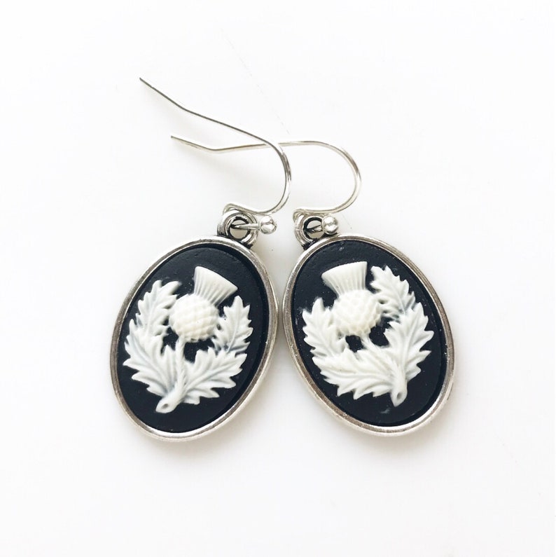 May include: A pair of silver dangle earrings with black oval-shaped settings and white enamel thistle designs.