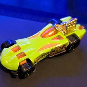 May include: A lime green toy race car with orange and gold accents. The car has black wheels and a gold engine detail. The car's design includes orange flame-like shapes and a detailed pattern on the side.