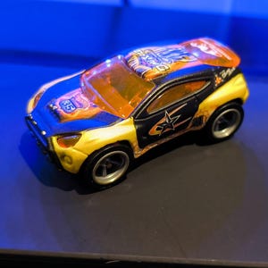 May include: A small, colorful toy car with a yellow and blue body, black accents, and orange tinted windows. The car features a star graphic and the number 65. This toy car is a collectible item.